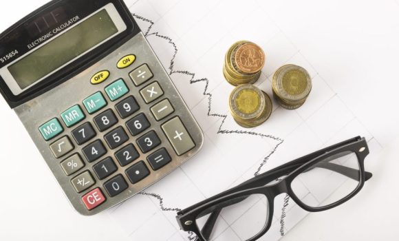 Calculadora plateada usada, gafas negras y monedas apiladas sobre un gráfico lineal, para explicar el cálculo del valor fiscal de una propiedad.